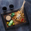 A wooden tray showcasing skewered grilled meat seasoned with The Spice Lab Ground Coriander, accompanied by sliced cucumber, lime halves, chili, cilantro, onions, and a bowl of dipping sauce. Ideal for BBQ restaurants or large foodservice gatherings—this generous 1 lb bulk spice helps elevate every dish. Two glasses of dark beverages sit above the tray on a dark surface.