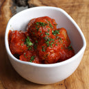 A white bowl filled with meatballs covered in tomato sauce, garnished with chopped parsley and infused with The Spice Lab Mediterranean Citrus Seasoning, sits on a wooden surface.