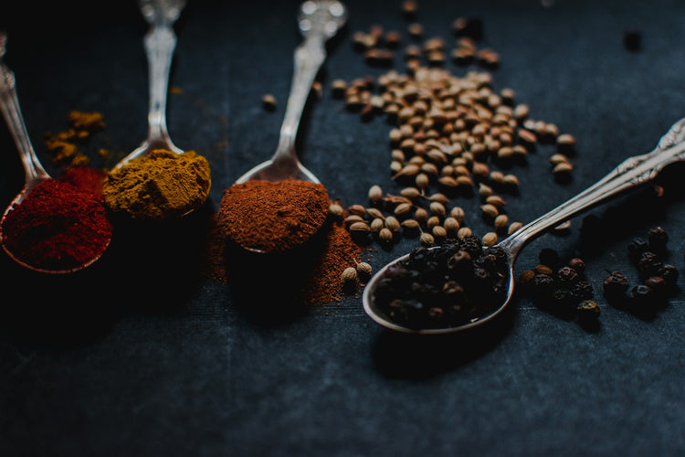 Four ornate silver spoons on a dark surface, each holding a different spice, including red, yellow, brown, and black powders. Loose peppercorns and coriander seeds are scattered nearby.