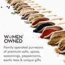 Several wooden scoops filled with vibrant spices and seeds—including The Spice Lab Himalayan Pink Salt Coarse Grain—are lined up diagonally on a white surface. Text reads: Women Owned. Family operated purveyors of premium salts, spices, seasonings, peppercorns, exotic teas & unique gifts—perfect for your salt grinder or gourmet kitchen from The Spice Lab.