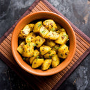 A clay bowl filled with Indian spiced potato cubes, seasoned with The Spice Lab Ground Coriander 1 lb Bulk Foodservice Spice and garnished with fresh coriander, placed on a bamboo mat over a dark textured surface.
