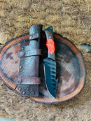 The Texan Knives Hunting Skinning Knife features a 10" J2 steel blade and a wangi/resin handle, resting on a wooden slab beside its brown leather sheath, all displayed on coarse natural fibers.