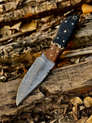 The Texan Knives Handmade 8" Damascus Skinning Knife features a high-carbon patterned blade and a buffalo horn and rosewood handle, displayed on a weathered log with dried leaves. Includes leather sheath.