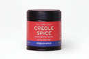 A round jar of World Spice Creole Spice handcrafted blend features a red label with white text outlining its flavor profile—Heat, Umami, Earthy—and the net weight (1.69oz / 48g). The design includes a blue band at the bottom displaying the World Spice brand name. Crafted as a classic Creole seasoning, this blend brings peppery heat and evokes the signature trinity mix found in Creole cuisine.
