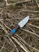 The Texan Knives Gutting Knife is a 10" handmade Damascus steel hunting knife featuring a patterned blade, rosewood or resin handle, and comes with a leather sheath, displayed on dry twigs and grass.