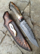 The Texan Knives Western Bowie Knife features a 15" hand-forged J2 steel blade, an antler horn or resin handle, and comes with a rustic leather sheath— all beautifully displayed on a stone surface.