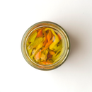 A jar of Taycte Famiglia Crispino Italian Mixed Vegetable Antipasto by Taycte—marinated garden vegetables in EVOO—is filled with vibrant yellow, orange, and green strips, creating a colorful display when viewed from above against a white background.