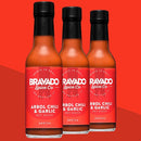 Three bottles of Bravado Spice Co Árbol Chili & Garlic Hot Sauce, each featuring red labels and black caps, are showcased against a matching red background.