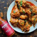 A plate of saucy chicken drumsticks garnished with cilantro sits beside a bottle of Bravado Spice Co Árbol Chili & Garlic Hot Sauce on a wooden table, ready to add a burst of bold Mexican hot sauce flavor to your meal.