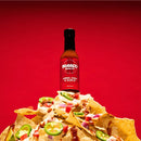 A bottle of Bravado Spice Co Árbol Chili & Garlic Hot Sauce sits atop a pile of nachos with cheese, jalapeños, onions, and sauce, set against a solid red background.