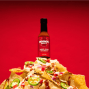 A bottle of Bravado Spice Árbol Chili & Garlic Hot Sauce from Bravado Spice stands behind nachos topped with jalapeños, onions, and a drizzle of sauce, set against a vivid red background. Made with authentic Chiles de Árbol for a bold medium-heat kick, this scene offers the promise of vibrant flavor in every bite.