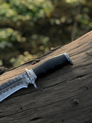 A close-up of the Texan Knives Best Bowie Knife—13" handmade Damascus steel blade with a resin horn handle and leather sheath—resting on a weathered log. The blurred greenery evokes an outdoor setting.