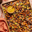 A sheet pan with roasted veggies, sliced sausage, and walnuts tossed in SALTOPIA Maple Mustard Seasoning Low Sodium: ANOTHER ONE BITES sits beside a bowl of mustard sauce and a partly visible orange napkin.