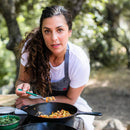 Chef Robin, in a white shirt and gray apron, cooks outdoors with a spoon over a skillet. Join her for the Messermeister HQ Cooking Class by Messermeister, enjoying pasta making amid lush green foliage.