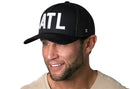 A smiling man wears the Haka Hat Atlanta White Block Trucker Hat (Black White ATL Block Snapback) by Haka Hat—a black cap with bold white ATL letters—paired with a light T-shirt, set against a white background.