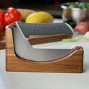 A curved NULU Knife by NULU Inc., featuring a textured gray handle and durable stainless steel blade, is displayed in its wooden Dock for convenient countertop storage. Fresh vegetables—including tomatoes and a lemon—are softly blurred in the background atop a white kitchen countertop.