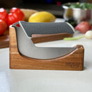 A curved kitchen knife with a gray handle is displayed in the NULU Knife Holder Dock by NULU Inc., crafted from acacia wood. The blurred background features fresh vegetables, such as tomatoes, on a kitchen counter, drawing attention to this elegant countertop knife storage solution.
