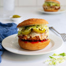 A crispy chicken sandwich with melted cheese, lettuce, pickles, and Terrapin Ridge Farms Red Pepper Jelly sits on a white plate. Another sandwich and utensils are blurred in the background.