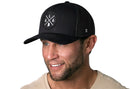A smiling man with a short beard wears a Haka Hat Alabama Trucker Hat | Black ALA X Snapback, featuring a circular logo with intersecting white lines and 'L A' on the front. He is dressed in a light-colored shirt.