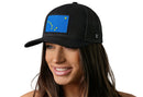 A woman with long brown hair smiles while wearing a Haka Hat Alaska Flag Trucker Hat— a black mesh snapback by Haka Hat featuring a blue patch with yellow stars forming the Big Dipper and Polaris of the Alaska state flag.