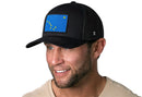 A man in a beige shirt wears the Haka Hat Alaska Flag Trucker Hat, a black mesh snapback by Haka Hat featuring a patch with eight yellow stars on a blue background. He is smiling and looking slightly to the side.