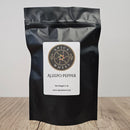 A black resealable pouch labeled Spice Wheel Aleepo Pepper 1/2 lb rests on a wooden surface. The packaging displays a round logo and product details in black and gold, emphasizing this gourmet pepper as perfectly crushed for culinary enthusiasts.