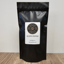 A black resealable pouch from Spice Wheel, labeled “Spice Wheel Aleepo Pepper 1 lb,” containing gourmet pepper flakes, stands upright on a wooden surface against a plain light background.