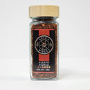 A glass jar of Spice Wheel Aleppo Pepper rests on a white background. The jar is sealed with a wooden lid and displays vibrant chili flakes known for their mild heat. Its label, adorned in black and red, features a circular logo, the brand name Spice Wheel, the product name Aleepo Pepper, and Net Wt. 58g.