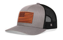 The Haka Hat American Flag Trucker Hat features a gray-black design with a brown leather patch, black mesh back, and USA snapback style.