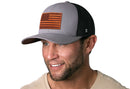 A man wears the Haka Hat American Flag Trucker Hat Leather in gray and black, featuring a leather patch with the USA flag and a black mesh back.