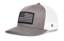 The Haka Hat American Flag Trucker Hat is a gray and white snapback by Haka Hat, featuring a black-and-gray American flag patch, mesh back, visible brim stitching, and a small logo tag on the side mesh.