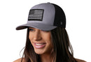 A woman with long brown hair smiles while wearing the Haka Hat American Flag Trucker Hat, a gray-black USA Tactical snapback by Haka Hat, against a clean white background.