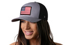 A woman with long brown hair smiles wearing the Haka Hat American Flag Trucker Hat in gray and black, featuring a USA embroidered flag patch on the front. The snapback stands out against the crisp white background.