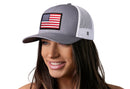A woman with long brown hair smiles and looks to the side while wearing the Haka Hat American Flag Trucker Hat, a gray-white USA snapback with a flag patch on the front.