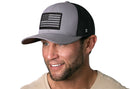 A man with a trimmed beard smiles in a light beige shirt, wearing the Haka Hat American Flag Trucker Hat—Gray-Black USA Tactical Snapback by Haka Hat, featuring a bold USA Tactical patch, set against a white background.