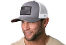 A man with a short beard smiles while wearing the Haka Hat American Flag Trucker Hat, a gray-white USA tactical snapback featuring a black and gray flag patch by Haka Hat, paired with a beige shirt.