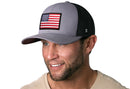 A smiling man wears a Haka Hat American Flag Trucker Hat, Gray-Black USA Snapback with an embroidered flag patch and a light beige shirt. The background is plain white.
