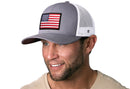 A man with a short beard and blue eyes smiles while wearing the Haka Hat American Flag Trucker Hat | Gray-White USA Snapback by Haka Hat, featuring an embroidered flag patch. He also wears a beige shirt.