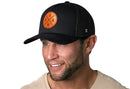 A man with a trimmed beard smiles, wearing the Haka Hat USA Trucker Hat Leather Black America Snapback featuring a brown circular USA and star emblem. He pairs it with a light shirt against a white background.