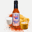 A bottle of Culture Hot Sauce American Dream from Culture Hot Sauce, featuring bold cayenne chili pepper flavor, stands alongside sliced corn and a cup of ranch dip topped with ranch seasoning, black pepper, and red seasoning powder on a white background.