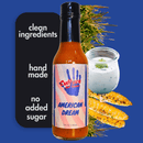 A bottle of Culture Hot Sauce American Dream by Culture Hot Sauce, labeled with "clean ingredients," "handmade," and "no added sugar." The background showcases grilled corn, fresh green herbs, and a jar of sauce on a blue surface. This American hot sauce is crafted with care to highlight bold flavors, featuring blends like cayenne chili pepper for an extra kick.