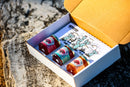 A white Brushfire Farms gift box labeled with the Three Amigos collection sits open on a rock, revealing three neatly arranged jars of jam—including signature pepper jam—and a vibrant illustrated card adorned with cacti and a sun. The box lid features the Brushfire Farms logo for an added personal touch.
