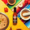 A bottle of Bravado Spice Ancho Masala Scorpion-Reaper Hot Sauce from Bravado Spice sits on a yellow surface surrounded by aromatic spices, tortillas, vibrant peppers, fresh garlic, and a colorful striped cloth.