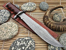 The Texan Knives Original 17" Antique Damascus Steel Bowie Knife with a rosewood handle and Damascus steel guard rests on its leather sheath, surrounded by speckled stones and a curved horn atop a bamboo mat.