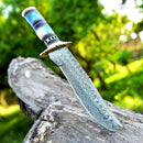 The Texan Knives Damascus Steel Antique Bowie Knife, 13" with brass guard and color bone & pakkawood handle, is displayed embedded in a weathered log with its handmade leather sheath nearby against a green outdoor background.