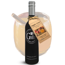 A dark bottle of Georgetown Olive Oil Co. Apple Cider White Balsamic Vinegar, expertly crafted in Modena, Italy, stands before a glass of apple cider garnished with a cinnamon stick. The bottle is accented by a gold tag featuring apple motifs and product details, emphasizing its distinguished White Balsamic Vinegar blend.
