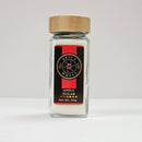 A clear glass jar with a wooden lid holds Spice Wheel Apple Sugar, made from granulated sugar mixed with dried apple powder. The front label displays the Spice Wheel brand name, the product name "Apple Sugar," and "Net Wt. 90g" in a black and red design featuring a windmill graphic.