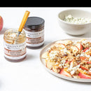 A plate of sliced apples, crumbled cheese, and walnuts sits beside a jar of Terrapin Ridge Farms Apple Maple Bacon Jam (5oz)—a perfect charcuterie board addition—with a spoon in the jar, another jam jar, and a bowl of cheese.