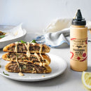 A plate holds two stacked halves of stuffed flatbreads filled with seasoned meat and drizzled with smoky sauce. Beside it sits Terrapin Ridge Farms Harissa Aioli Squeeze by Terrapin Ridge Farms, the perfect burger condiment.