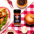 A jar of Bravado Spice Co Garlic & Árbol Seasoning by Bravado Spice Co sits on a checkered tablecloth, surrounded by an onion, garlic, black pepper, dried chilies, a spice bowl, and a sandwich filled with grilled chicken and vegetables—making it the perfect Tex-Mex seasoning to add bold flavor to any meal.